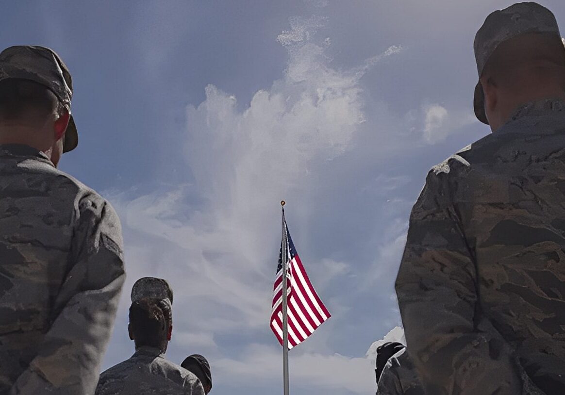 Soldiers facing American flag under cloudy sky.